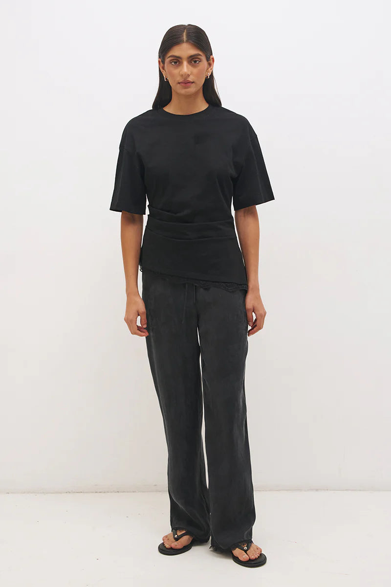 Mon Renn Liora T-Shirt in Black — full length front view showing crew neckline, cinched waist seaming and angled lace hem with side splits, styled with dark wide-leg trousers