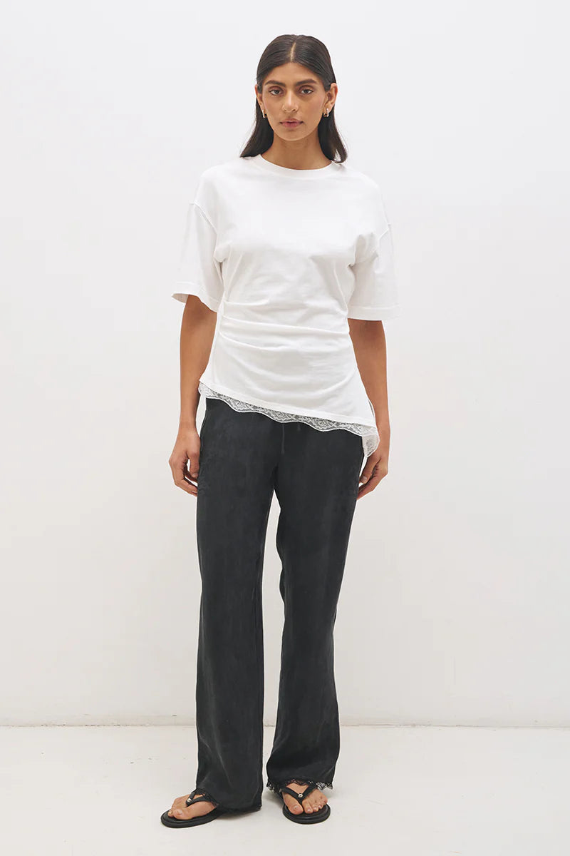 Mon Renn Liora T-Shirt in Off White — full length front view showing crew neckline, cinched waist seaming and angled lace hem with side splits, styled with dark wide-leg trousers